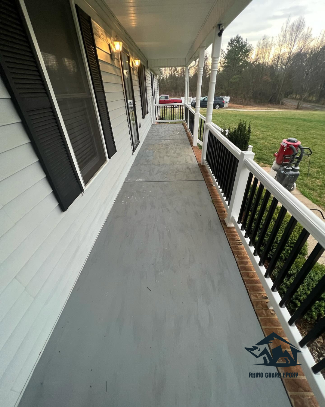 Long covered front porch with a gray epoxy floor, white siding, black shutters, and white railing overlooking the lawn.
