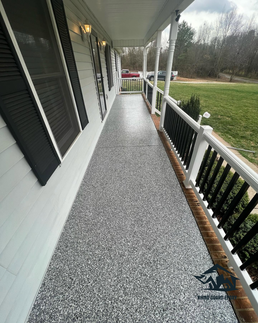 Long covered porch with a speckled gray epoxy floor, white railings with black balusters, and exterior lights along the wall.
