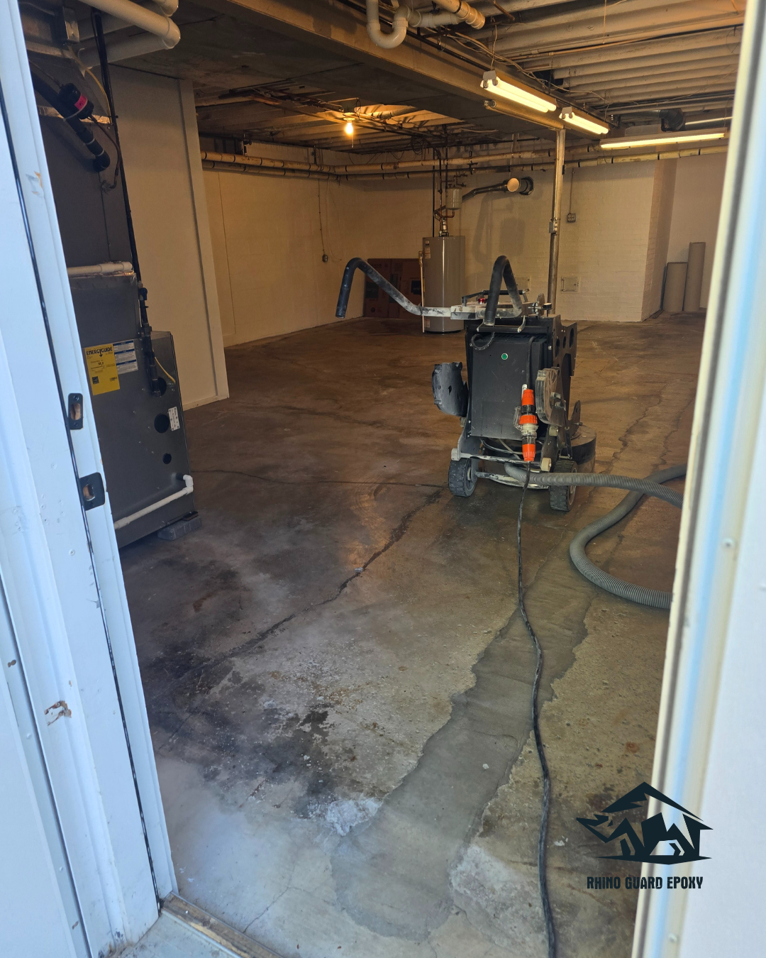 Basement workshop with an epoxy-coated concrete floor and a floor-applied machine connected to hoses; overhead pipes and fluorescent lights visible, Rhino Guard Epoxy logo in corner.