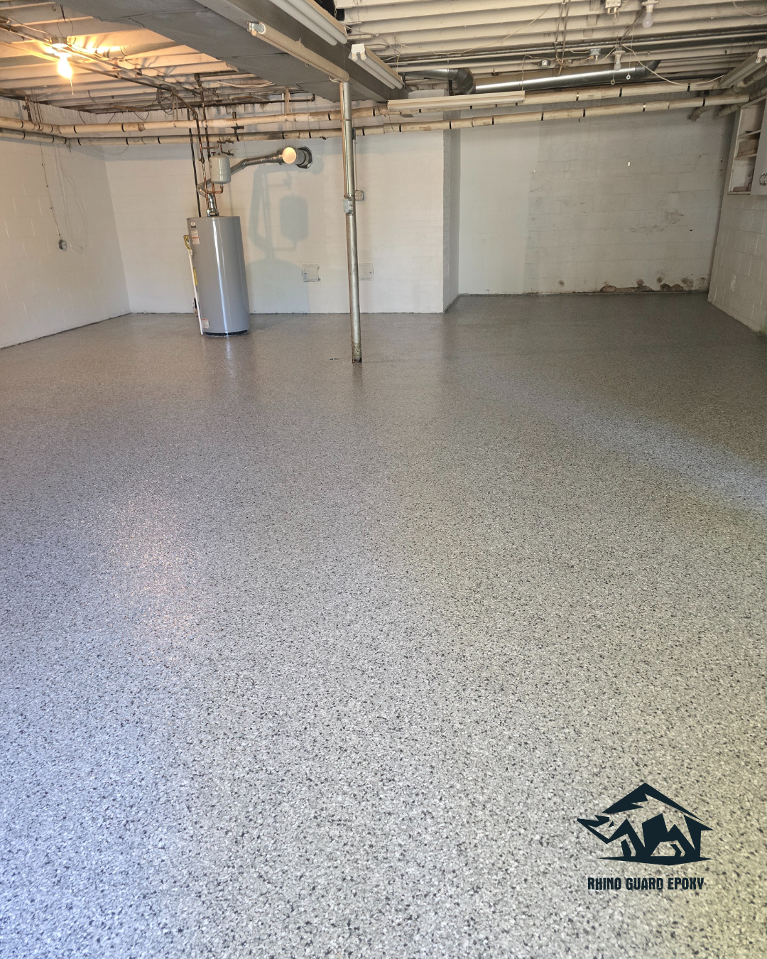 Basement with gray speckled epoxy floor, exposed pipes and water heater on the left, a central support post, white walls, and a Rhino Guard Epoxy logo bottom-right.
