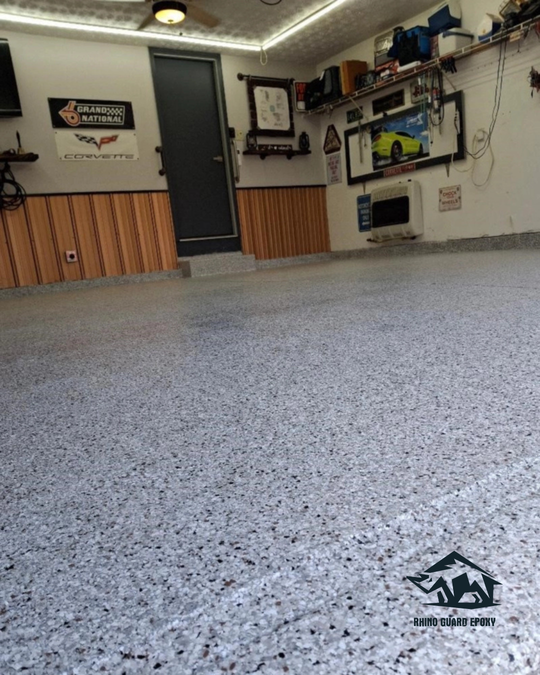 Indoor garage with a speckled gray epoxy floor, wood wainscoting, wall decor, and a Rhino Guard Epoxy logo in the lower right corner