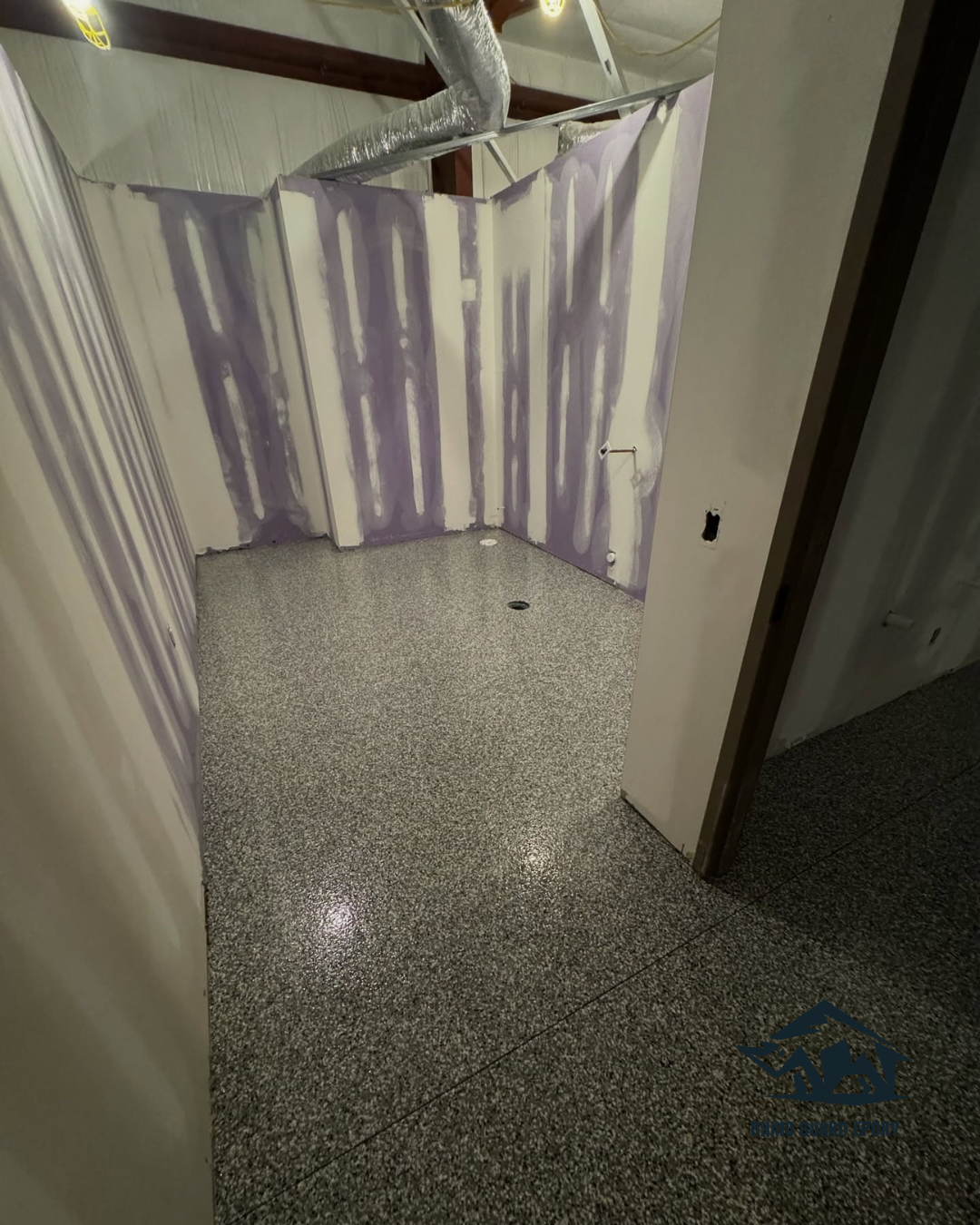 Construction site bathroom with partially finished purple drywall mud on studs, exposed ducts overhead, and a speckled tile floor. Watermark logo in bottom right corner.