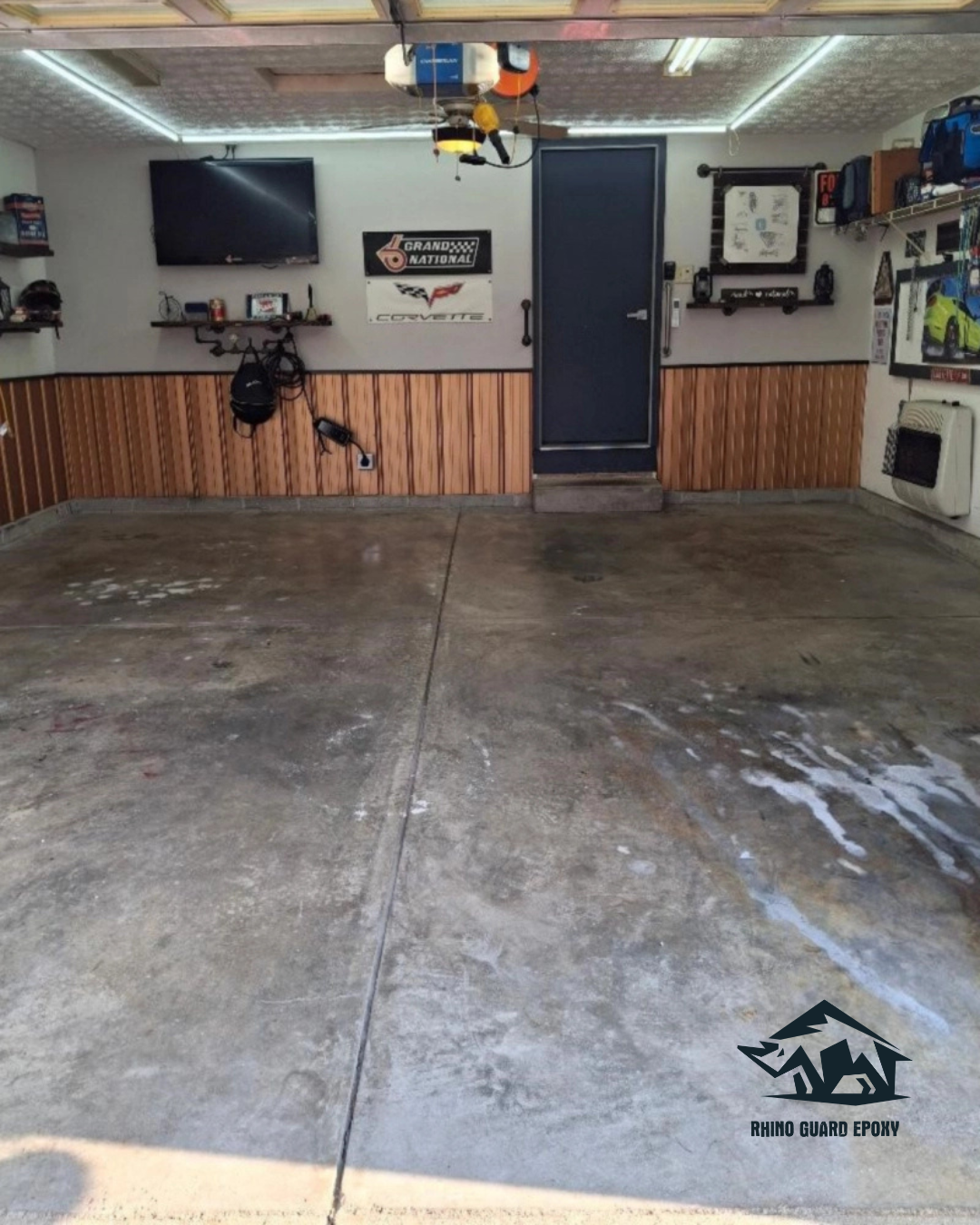 Tidy garage with a grey epoxy concrete floor, wooden wainscoting, blue door in the center, wall shelves and tools, and a Rhino Guard Epoxy logo in the lower right corner.