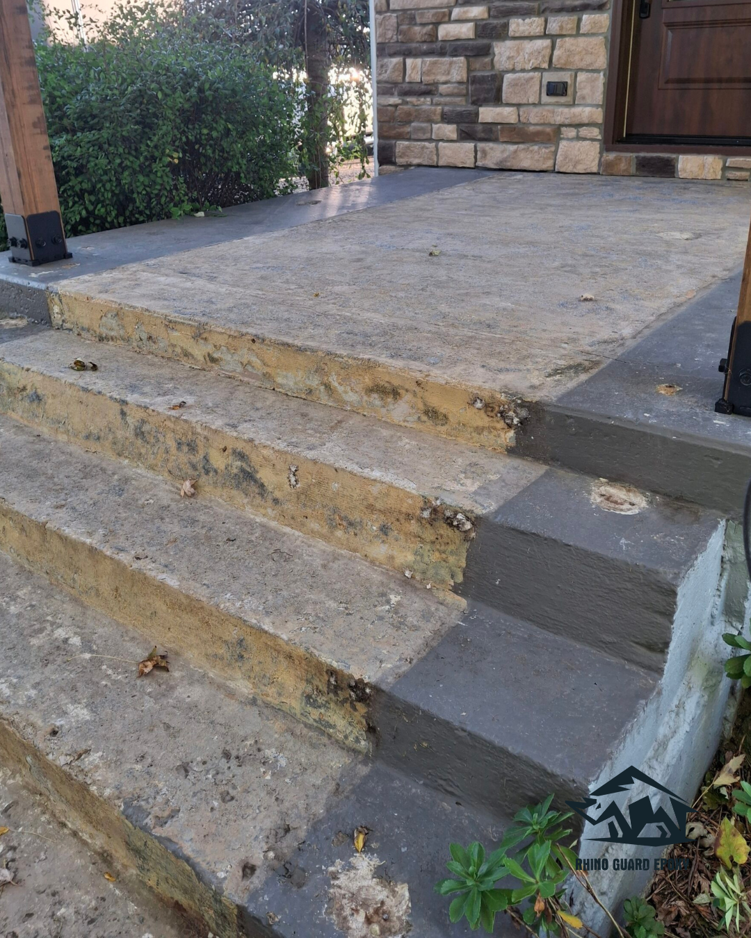 Worn concrete front steps with chipped edges leading up to a stone-clad entryway door, flanked by wooden posts and greenery nearby.
