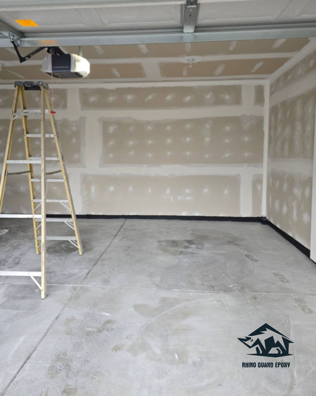 Empty garage under renovation with a tall ladder, a ceiling-mounted garage door opener, and unfinished drywall panels; logo reads 'Rhino Guard Epoxy' in the corner.