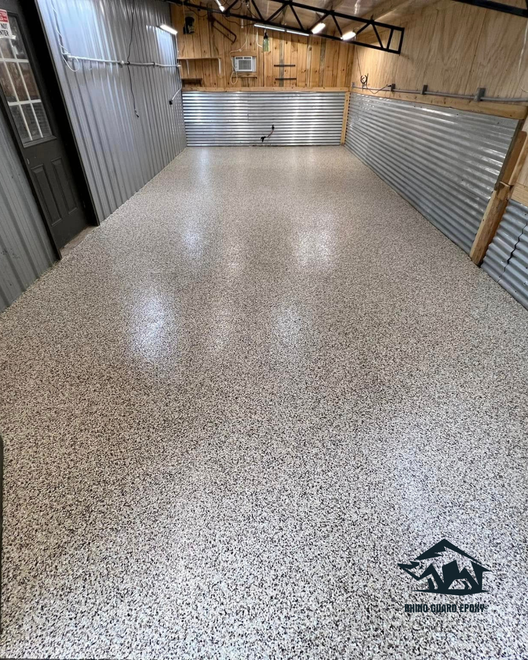 Empty garage workspace with speckled epoxy floor, corrugated metal walls, and wooden upper walls under bright overhead lighting.