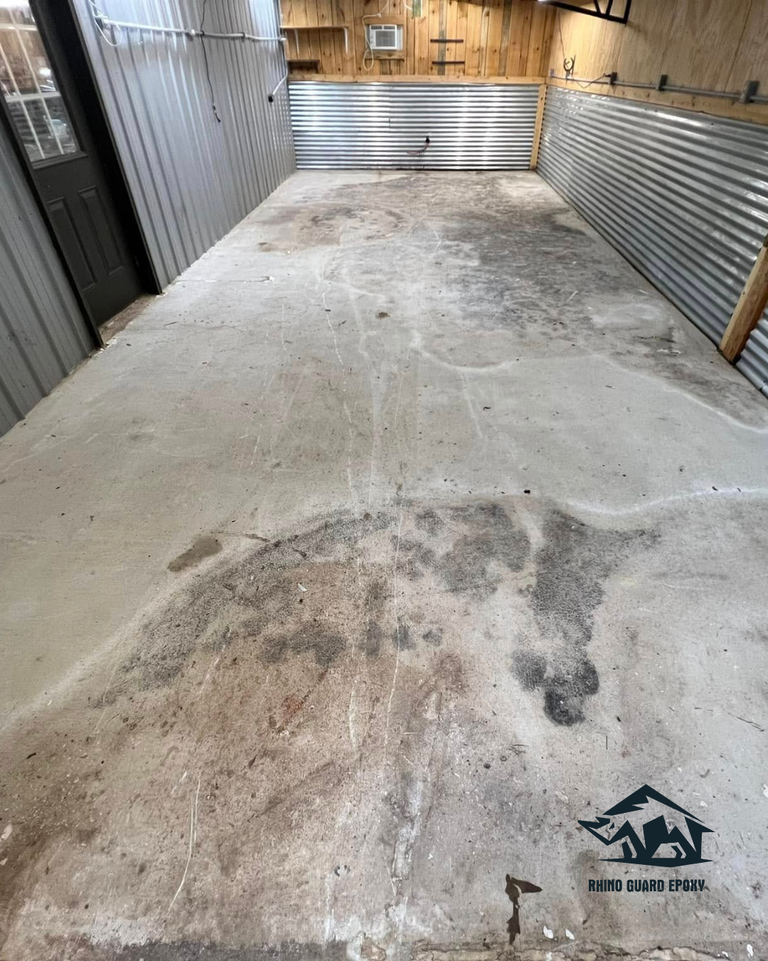 Empty metal-walled garage or storage unit with a stained concrete floor and wooden back wall, awaiting epoxy coating (logo bottom-right).