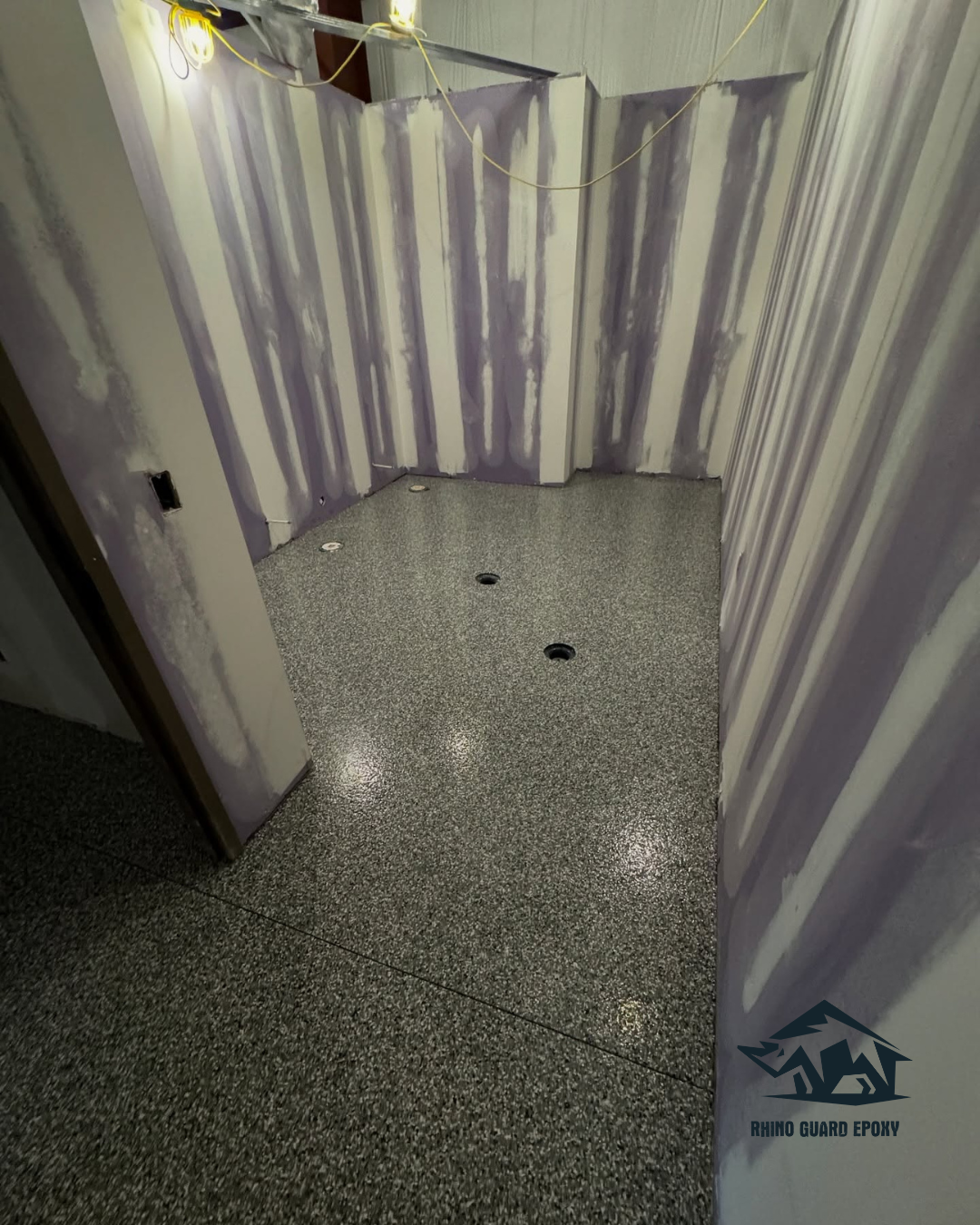 Unfinished bathroom with a speckled gray epoxy floor, striped drywall patches on the walls, and exposed wiring overhead; several circular floor holes visible.
