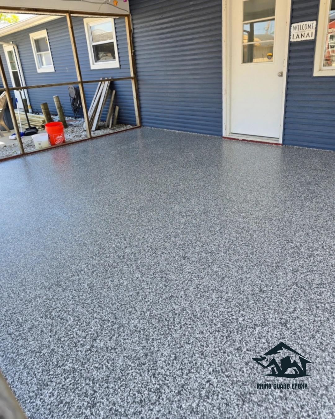 Blue exterior building with a white door and a speckled gray epoxy-coated porch floor; construction materials and an orange bucket visible in the background.