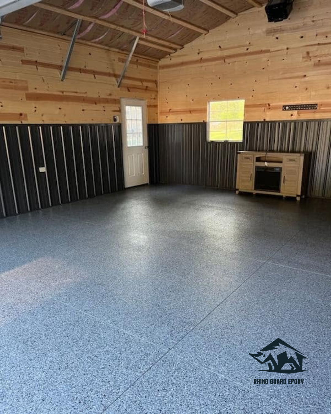 Interior of a workshop/garage with blue-speckled epoxy floor, wood-paneled walls and metal wainscoting, exposed ceiling beams, a white door, a small window, and a wooden cabinet with a small oven in the corner; Rhino Guard Epoxy logo in the bottom-right corner.